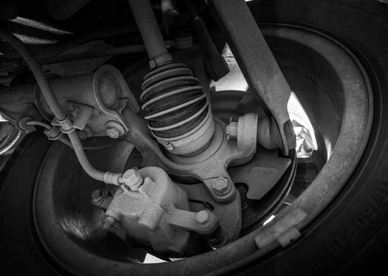 Monochrome close up view of a vehicle suspension assembly including CV axle control arm and steering components, photographed underneath a car inside an automotive repair workshop.